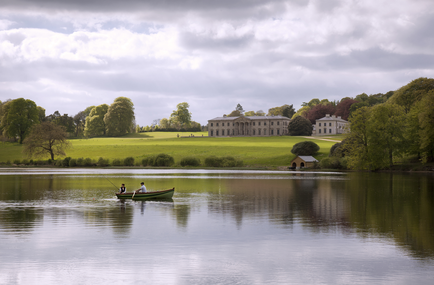 Two people fishing from a rowboat on a lake with a large house in the background.