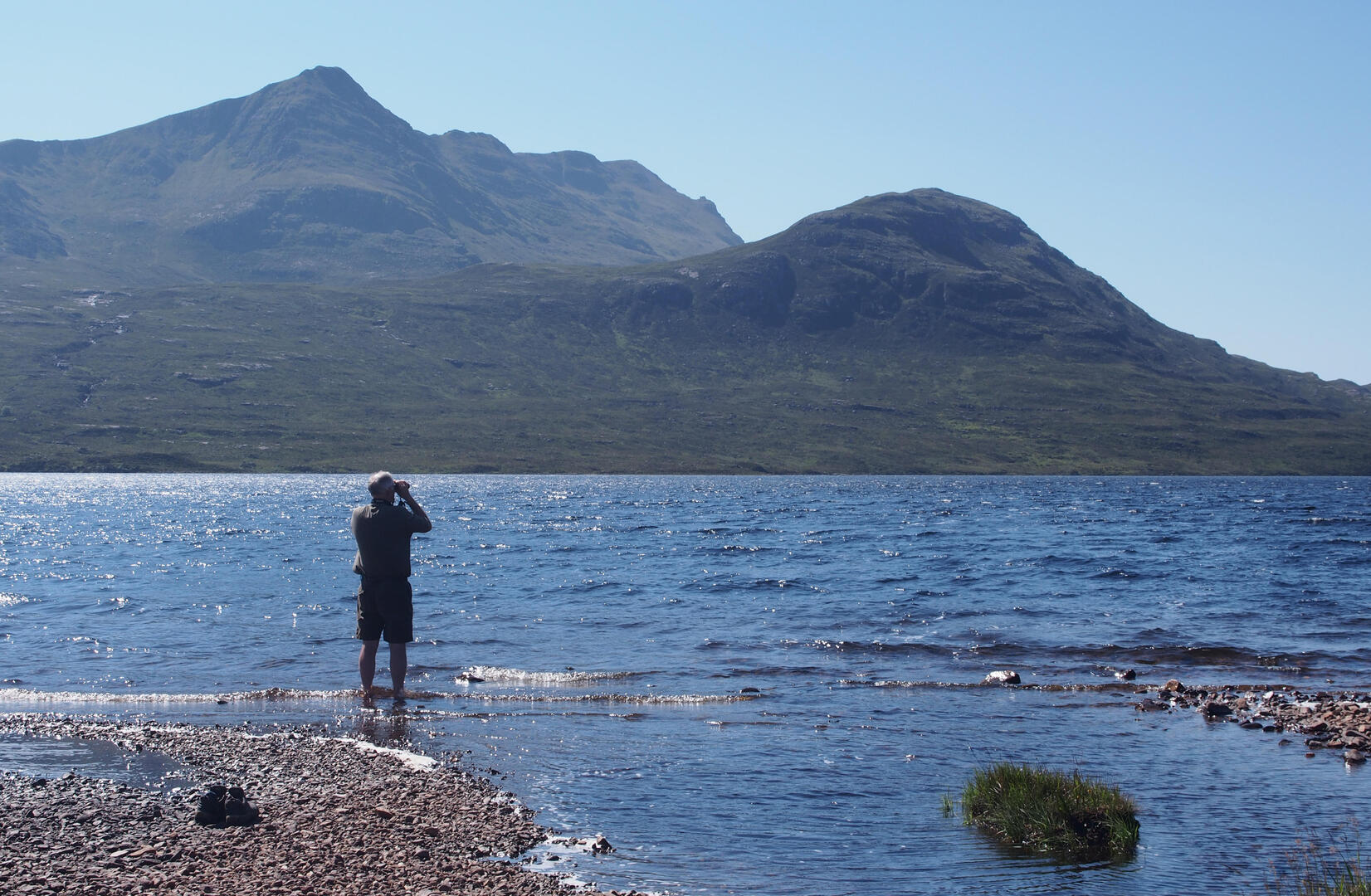 Man using binoculars by a lake with mountains in the background.
